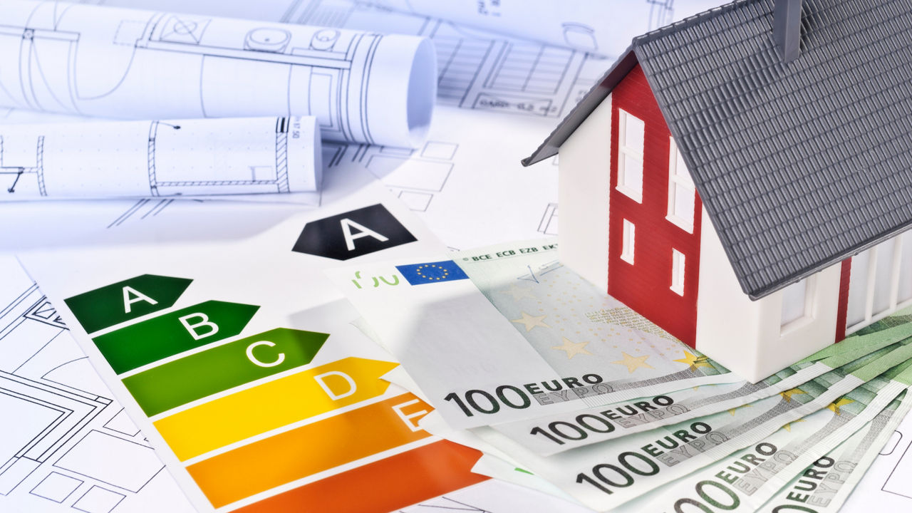 Modellhaus auf Euro-Scheinen neben einer farbigen Energieeffizienzskala von A bis F, im Hintergrund Baupläne.