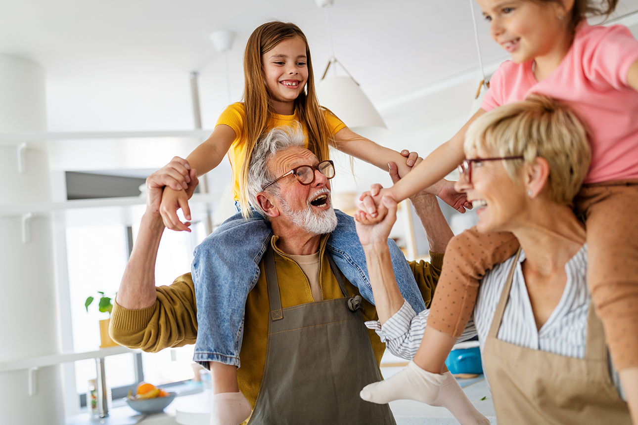 Auf dem Bild sind Oma und Opa zu sehen, die ihre Enkelkinder auf den Schultern tragen und gemeinsam Zeit verbringen.