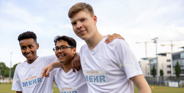 Drei Jugendliche in weißen Trikots stehen Arm in Arm nebeneinander und lächeln. 