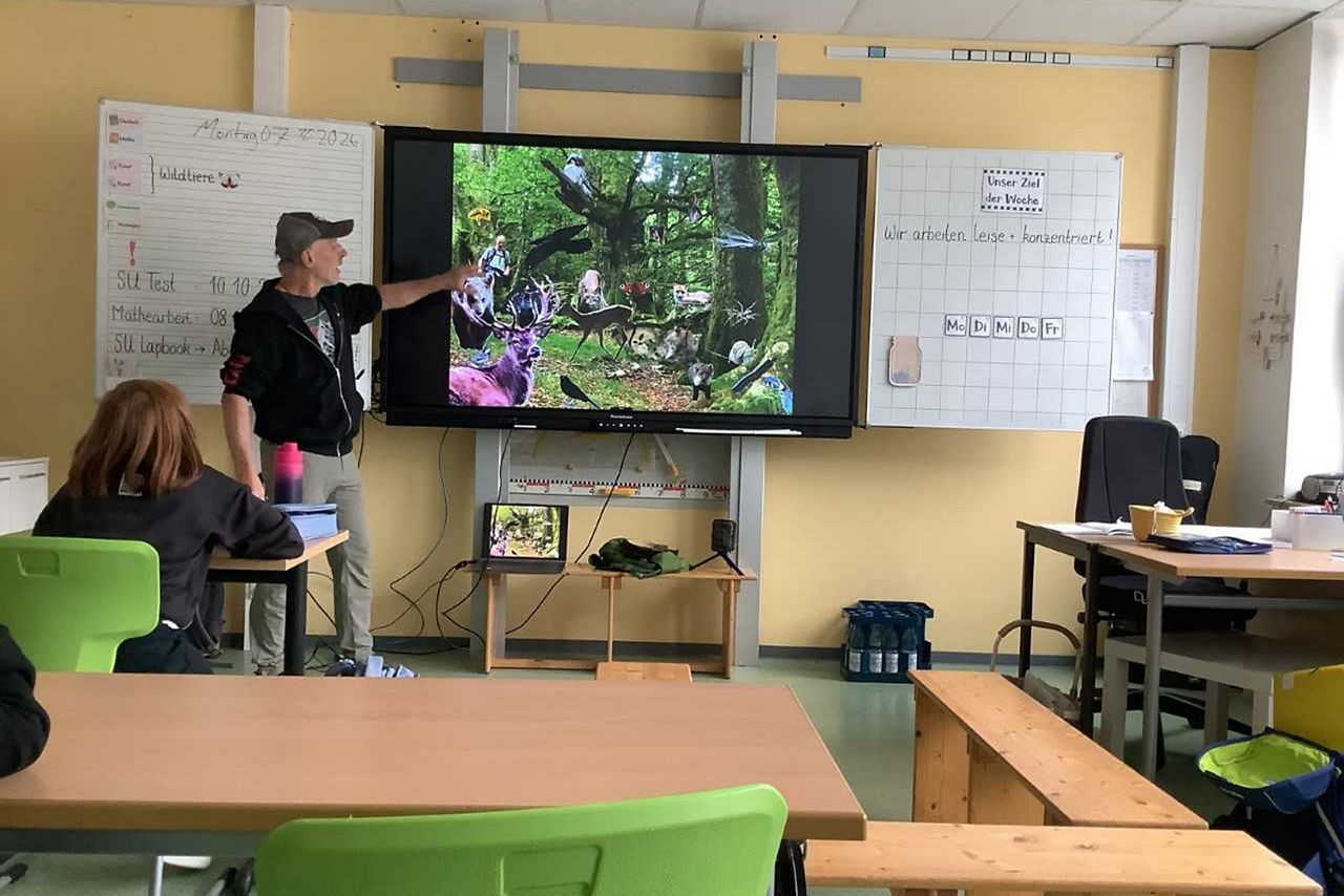 Ein Referent zeigt in einem Klassenzimmer auf ein großes Bildschirmbild mit verschiedenen Wildtieren im Wald.