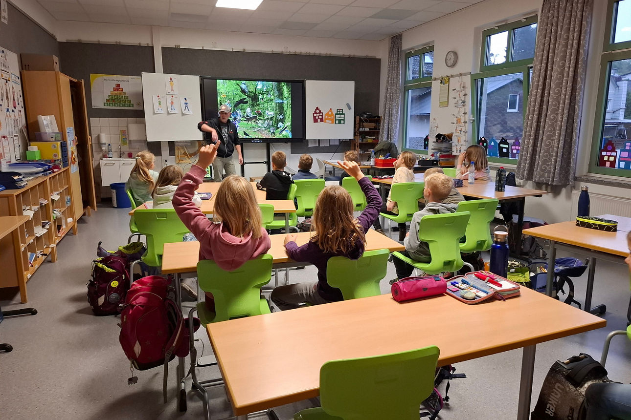 Grundschulkinder sitzen in einem Klassenzimmer mit grünen Stühlen und hören einem Vortrag über Wildtiere zu. 
