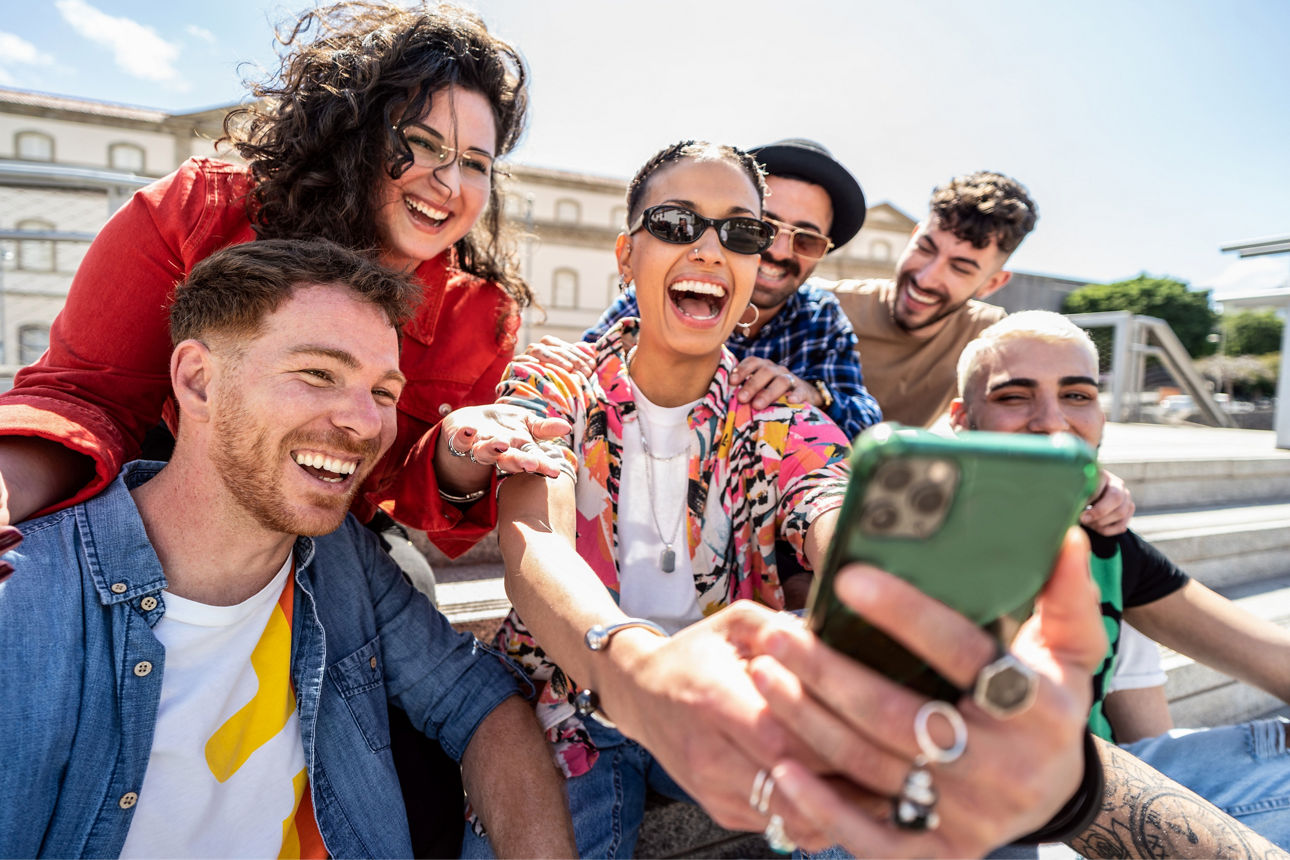 Sechs junge Erwachsene lachen und machen gemeinsam ein Selfie bei sonnigem Wetter