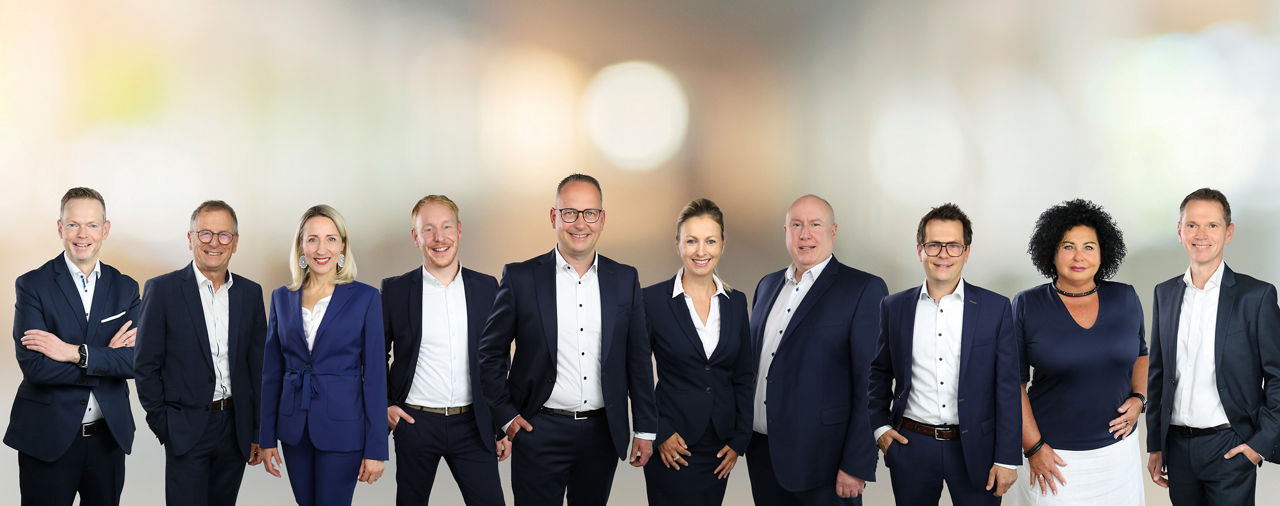 Eine Gruppe von elf Personen in Businesskleidung steht nebeneinander vor einem unscharfen Hintergrund.