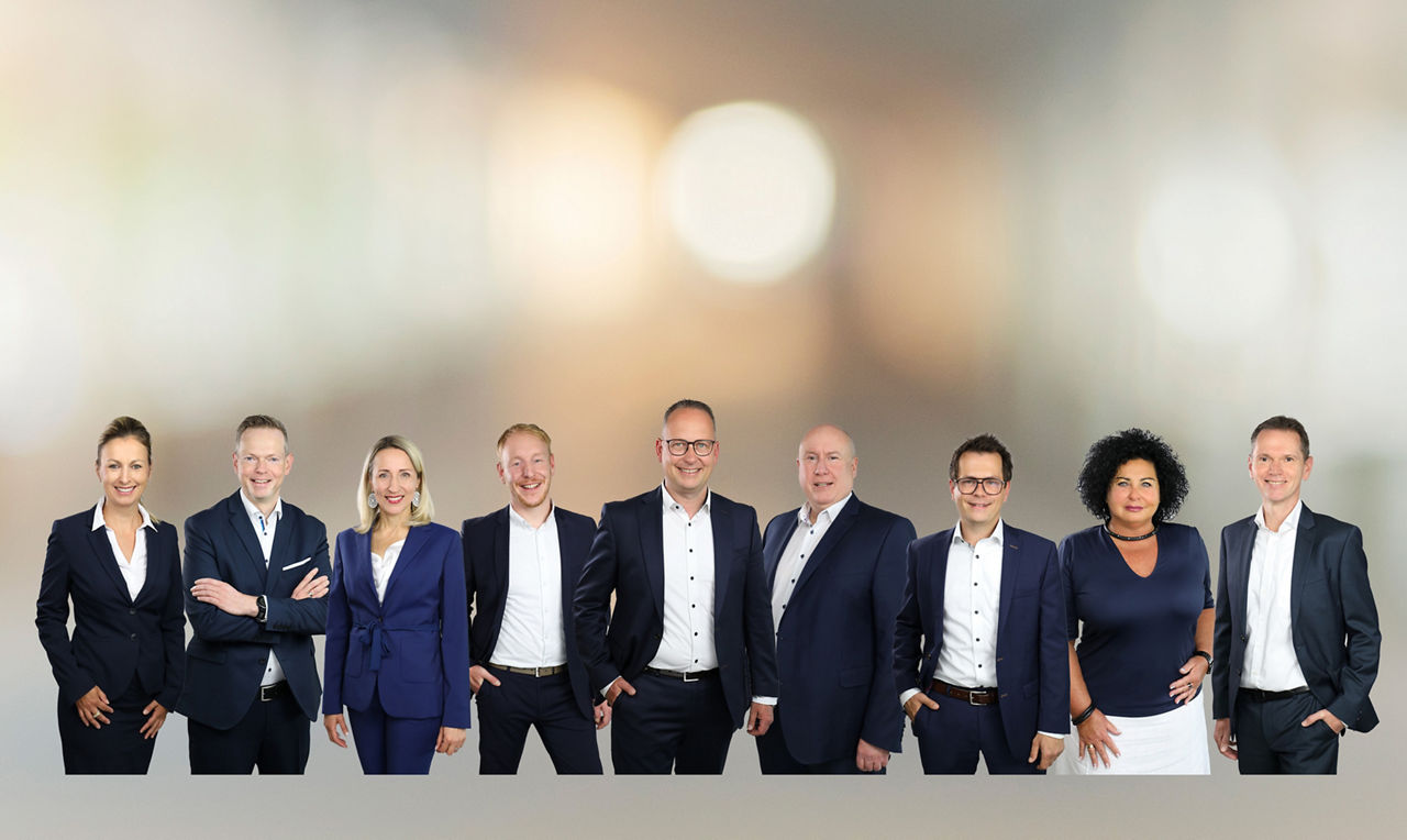 Eine Gruppe von elf Personen in Businesskleidung steht nebeneinander vor einem unscharfen Hintergrund.