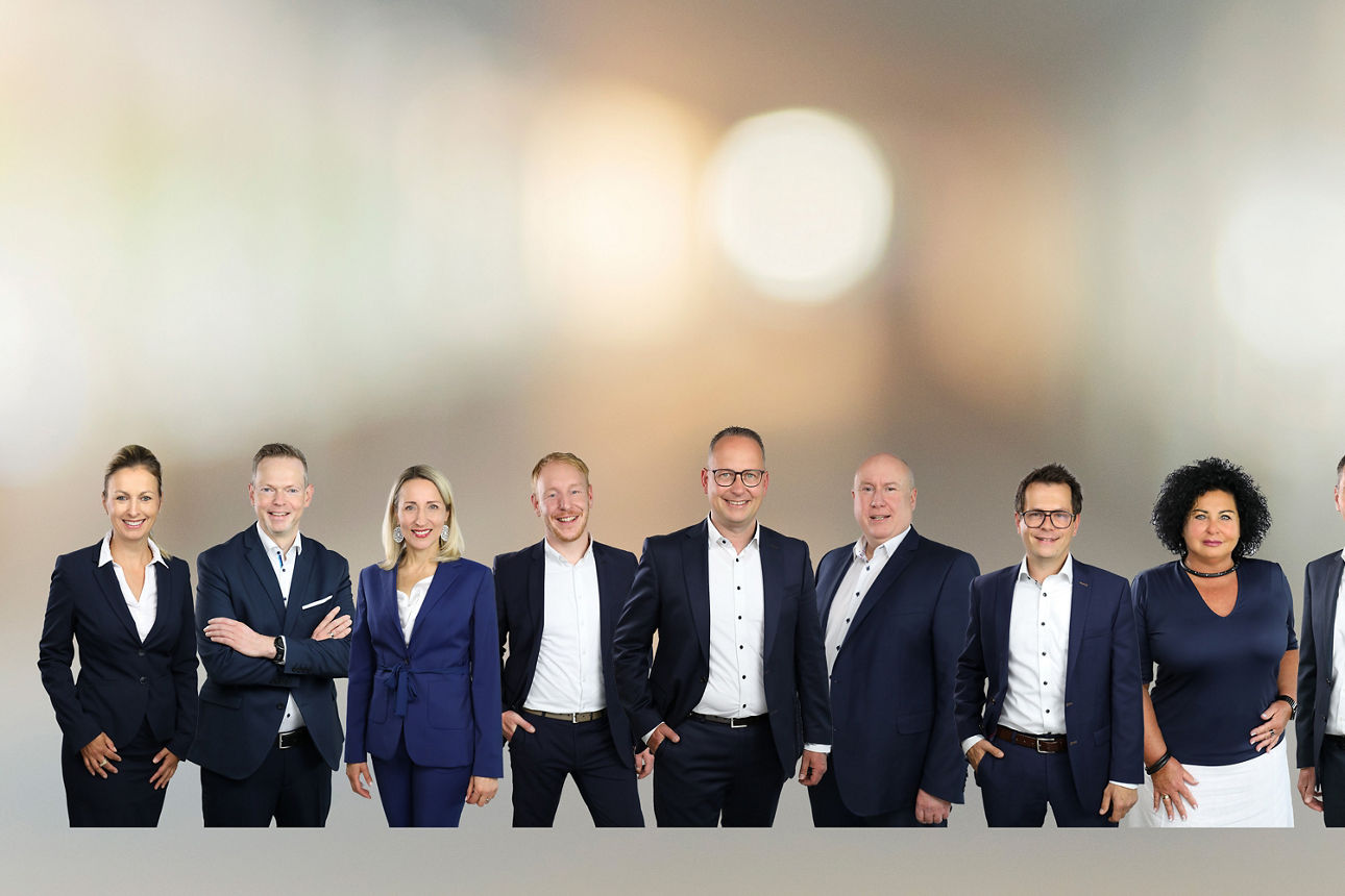 Eine Gruppe von neun Personen in Businesskleidung steht nebeneinander vor einem unscharfen Hintergrund.