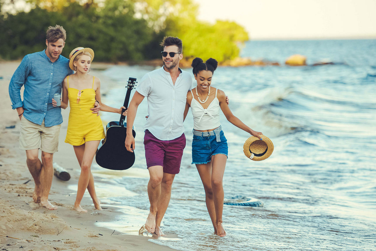 Zwei Pärchen gehen am Strand mit Gitarre und Sonnenhut spazieren