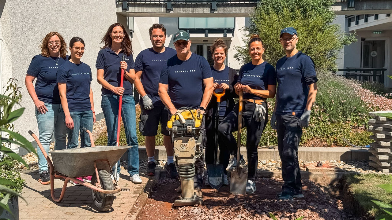 Team bei ehrenamtlichem Garteneinsatz, mit Werkzeug und Schubkarre, vor einem Gebäude bei sonnigem Wetter.
