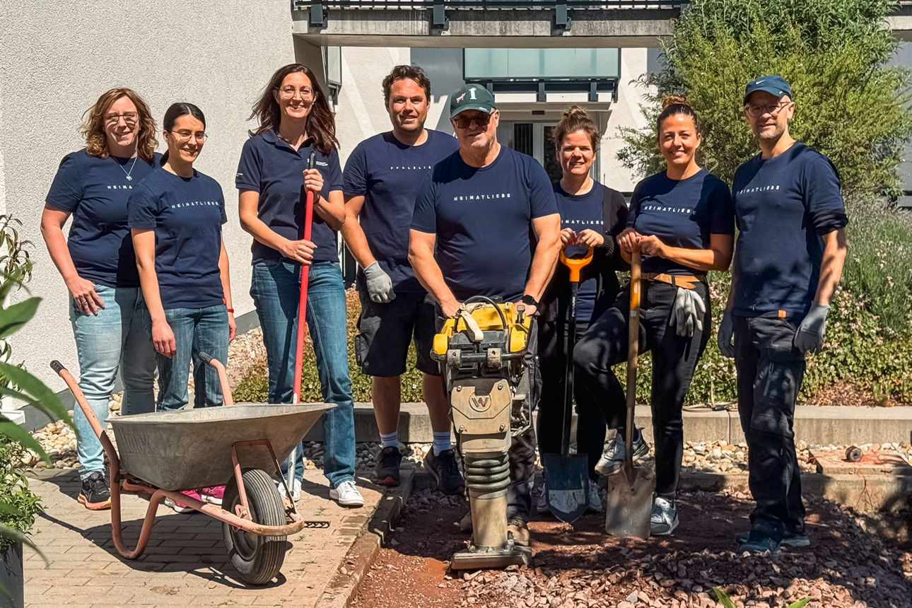 Team bei ehrenamtlichem Garteneinsatz, mit Werkzeug und Schubkarre, vor einem Gebäude bei sonnigem Wetter.