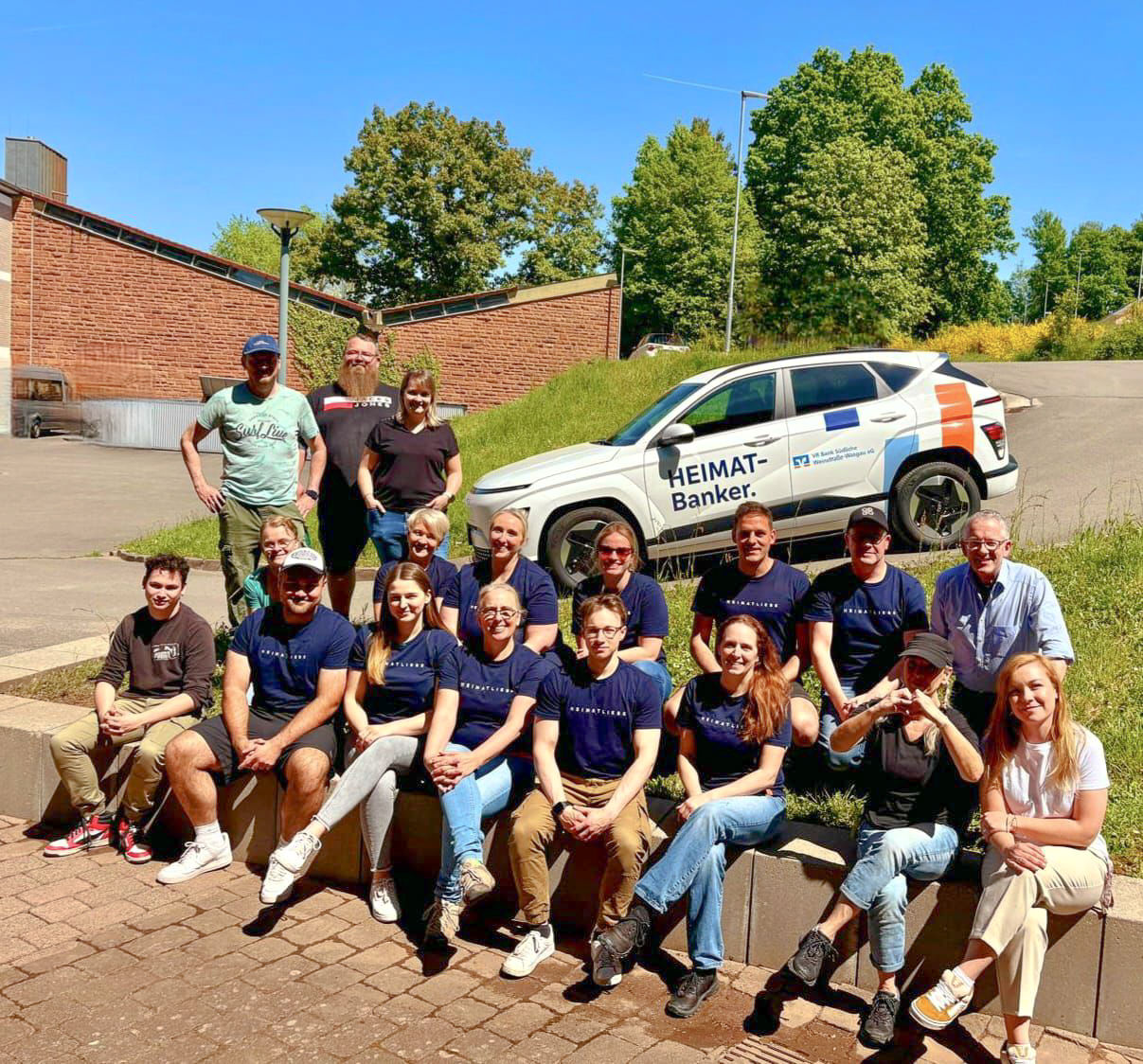 Gruppe von Mitarbeitenden sitzt und steht vor einem Auto mit „HEIMAT-Banker“-Aufdruck an sonnigem Tag im Freien.
