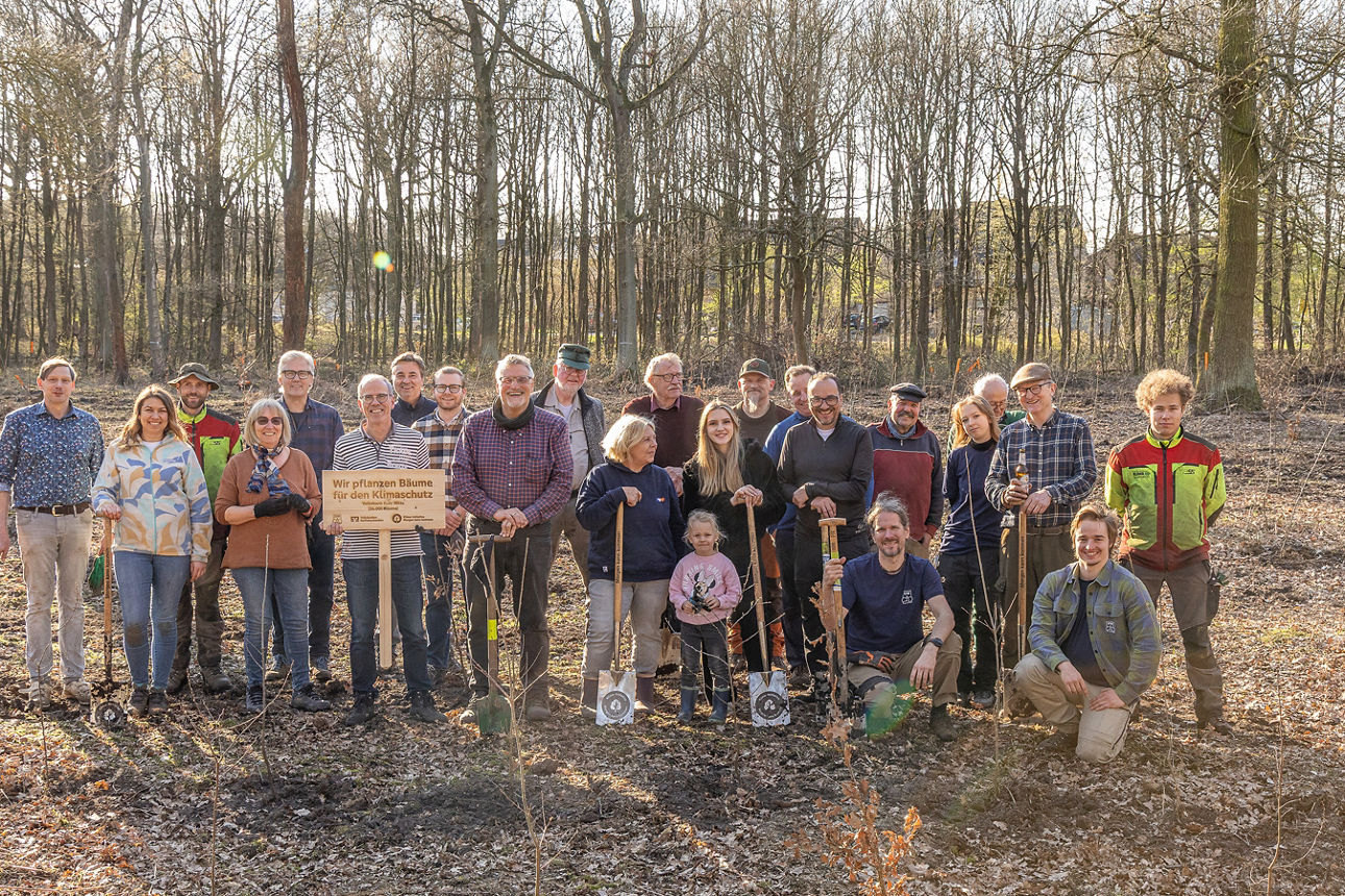 Eine große Gruppe Menschen steht gemeinsam in einem offenen Waldgebiet und posiert für ein Gruppenfoto. Einige halten Spaten oder Setzlinge in der Hand, andere ein Holzschild. Die Umgebung zeigt einen sonnigen Tag mit langen Baumstämmen im Hintergrund. Das Bild vermittelt den Abschluss einer gemeinsamen Pflanzaktion. Aufgenommen im Rahmen der Baumpflanzung der Volksbank Ruhr Mitte am 18.03.2026