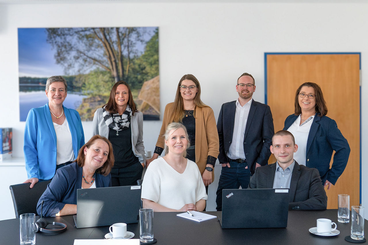Drei Personen sitzen nebeneinander vor Laptops und fünf Personen stehen dahinter 