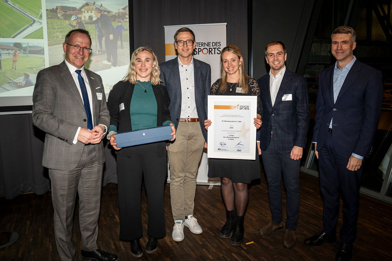 Fünf Personen stehen bei einer Preisverleihung zusammen und präsentieren Urkunde und Auszeichnung vor einer Bühne.