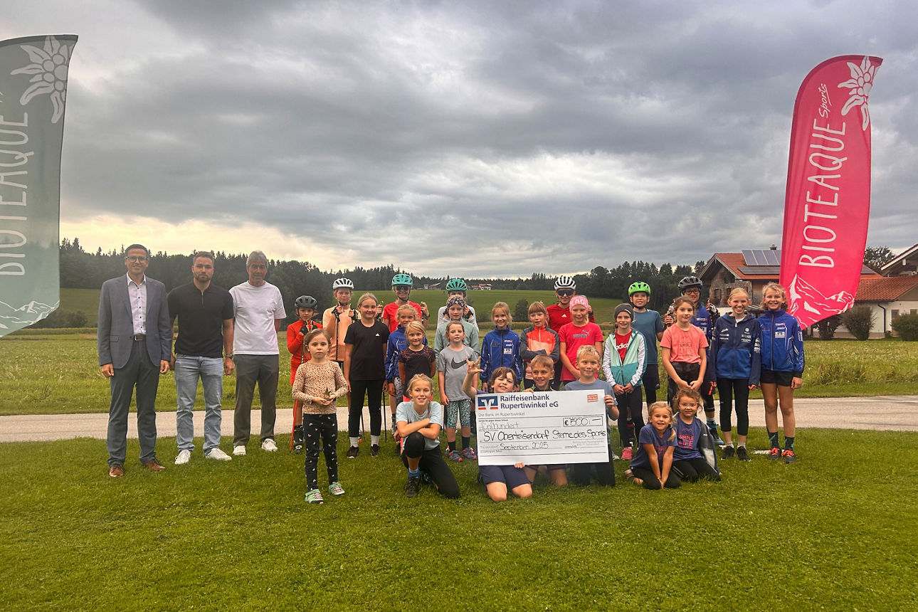 Große Gruppe von Kindern und Erwachsenen posiert auf einem Sportplatz und präsentiert gemeinsam einen Spendenscheck.