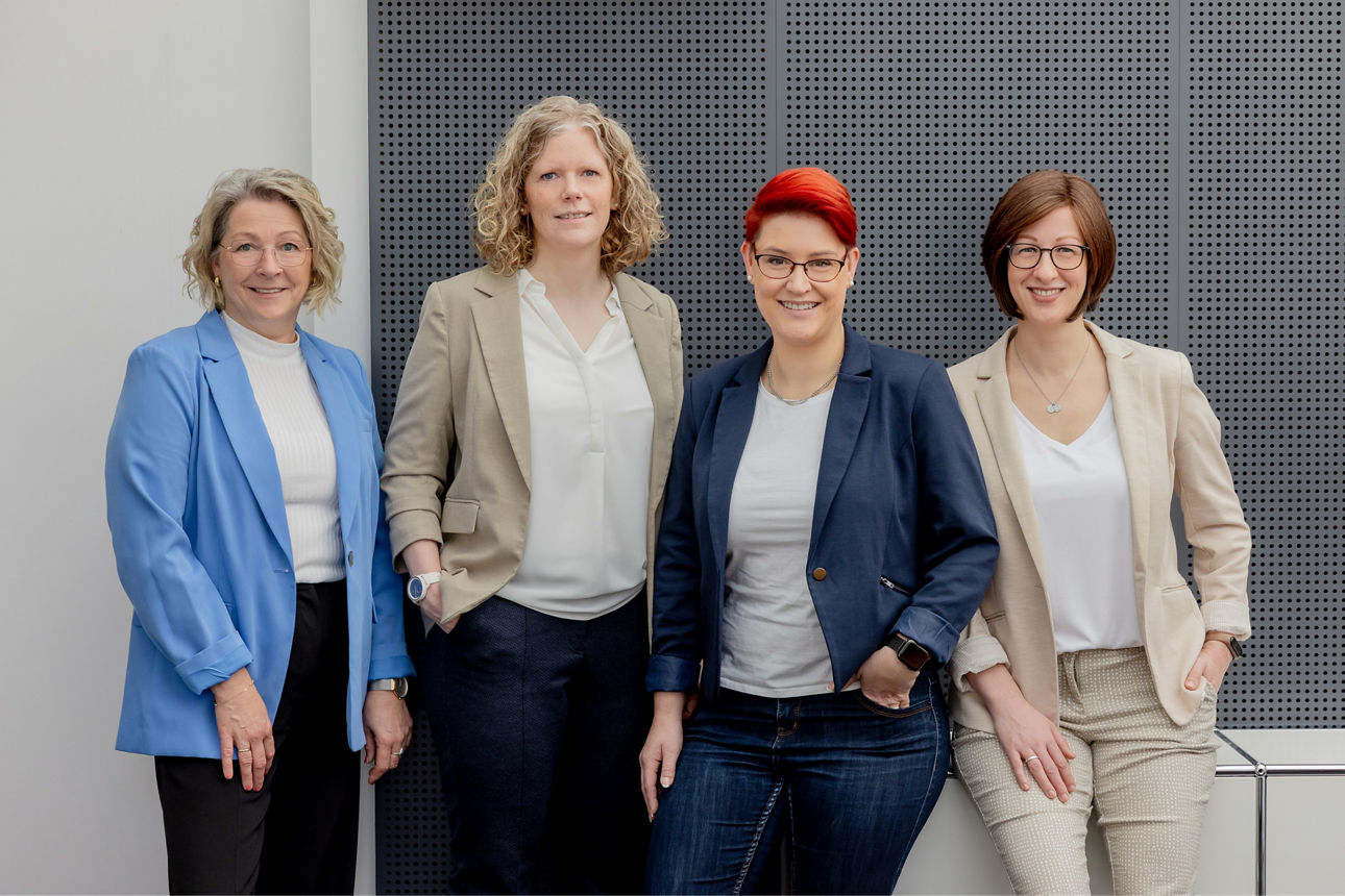 Vier Frauen stehen nebeneinander und lächeln in die Kamera. Sie tragen moderne Businesskleidung in Blau-, Beige- und Grautöne