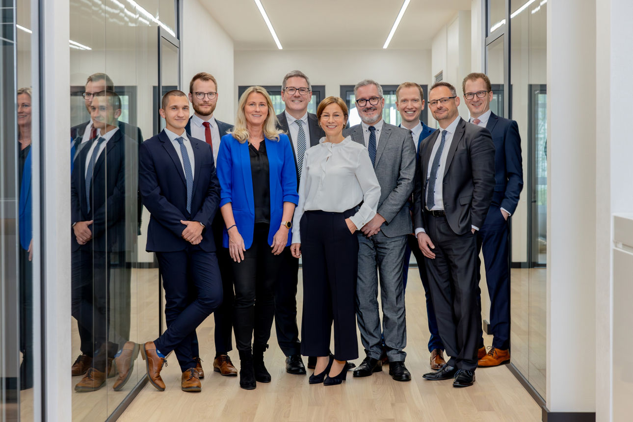 Eine Gruppe von Geschäftsleuten in formeller Kleidung steht in einem modernen Büroflur und lächelt in die Kamera.