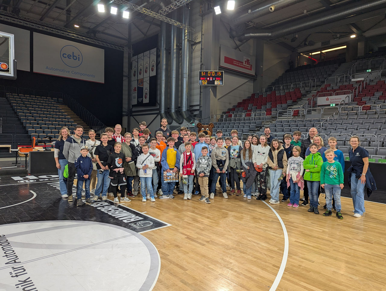 Auf dem Bild ist eine große Gruppe von Kindern und Erwachsenen auf dem Basketballfeld in einer Sporthalle versammelt. 
