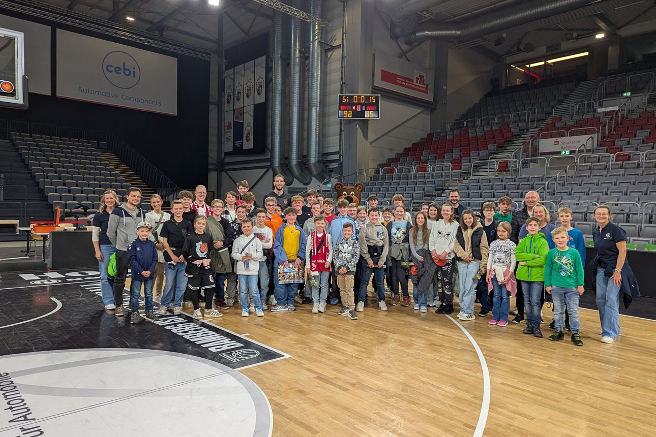 Auf dem Bild ist eine große Gruppe von Kindern und Erwachsenen auf dem Basketballfeld in einer Sporthalle versammelt. 