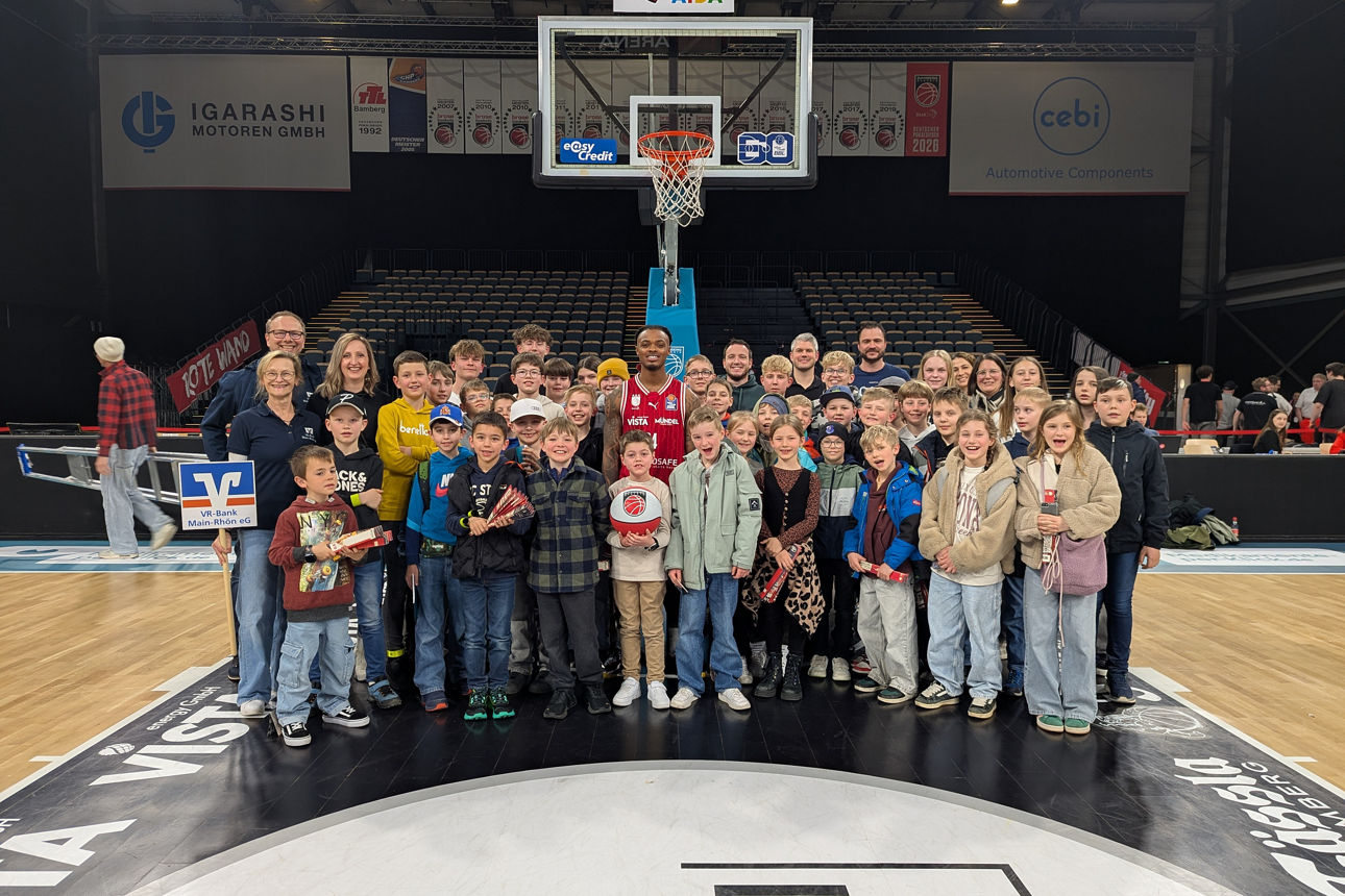 Das Bild zeigt 50 Kinder und Jugendliche des MeinClubs mit dem Betreuerteam der VR-Bank Main-Rhön sowie Cobe Williams, Bundesligaspieler der Bamberg Baskets, in der Mitte. Das Foto wurde in der Bose Arena auf dem Spielfeld unter einem Korb gemacht. 