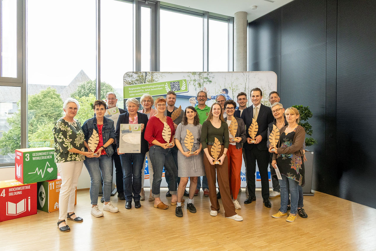 Auf dem Gruppenbild sind alle Gewinner des Klimapreises 2024 der Volksbank Ruhr Mitte zu sehen. Unter den 17 Personen befindet sich auch unser Vorstand Ingo Abrahams. Die Gewinner halten jeweils Trophähen in Form eines Holzblattes oder einer Urkunde in den Händen. Alle lächeln fröhlich in die Kamera.