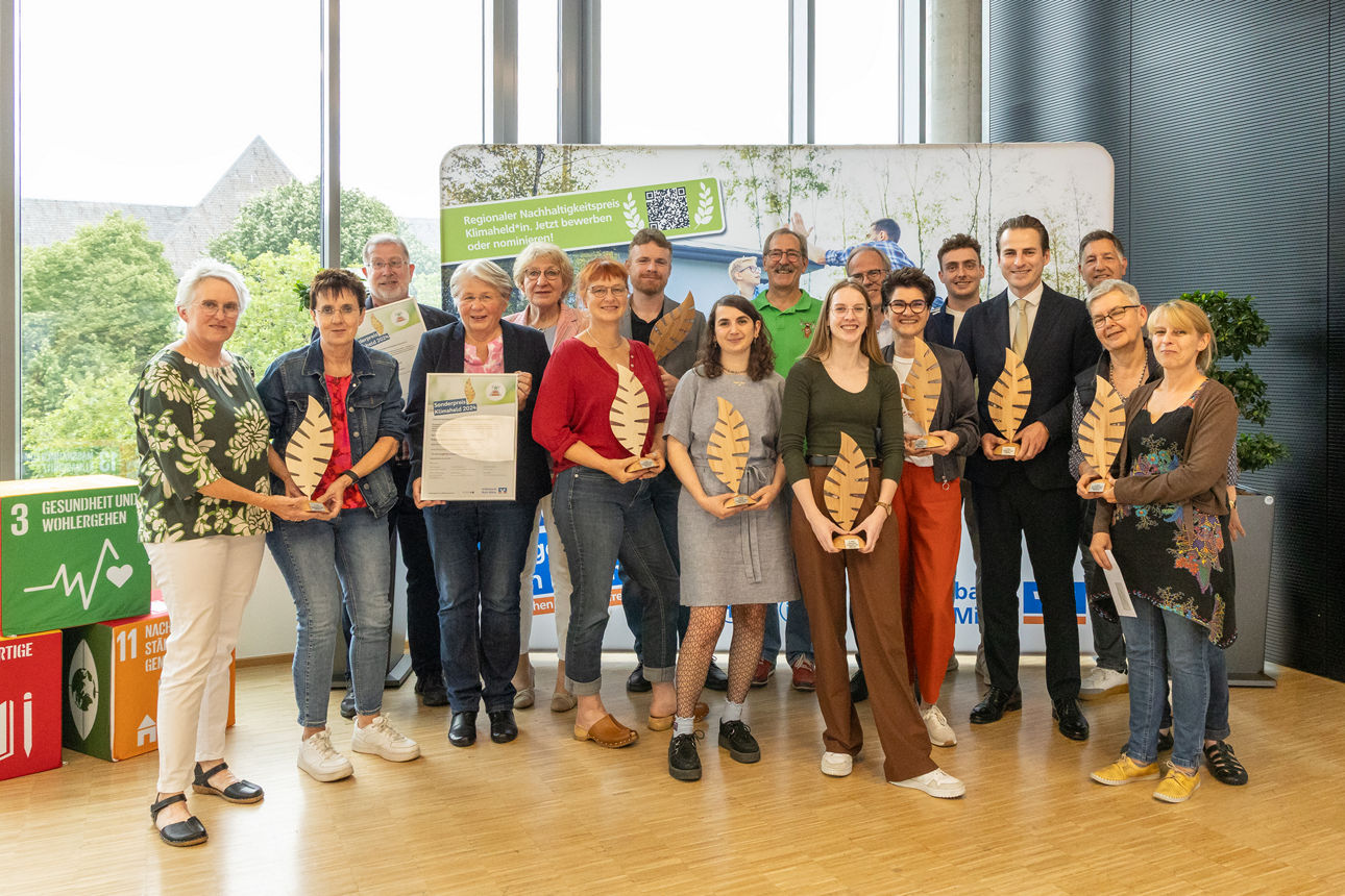 Auf dem Gruppenbild sind alle Gewinner des Klimapreises 2024 der Volksbank Ruhr Mitte zu sehen. Unter den 17 Personen befindet sich auch unser Vorstand Ingo Abrahams. Die Gewinner halten jeweils Trophähen in Form eines Holzblattes oder einer Urkunde in den Händen. Alle lächeln fröhlich in die Kamera.