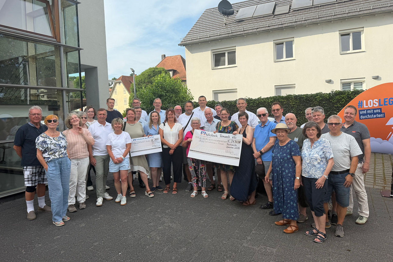 Eine Gruppe von Menschen versammelt sich im Freien zu einem Foto und hält dabei zwei feierliche Spendenschecks in den Händen
