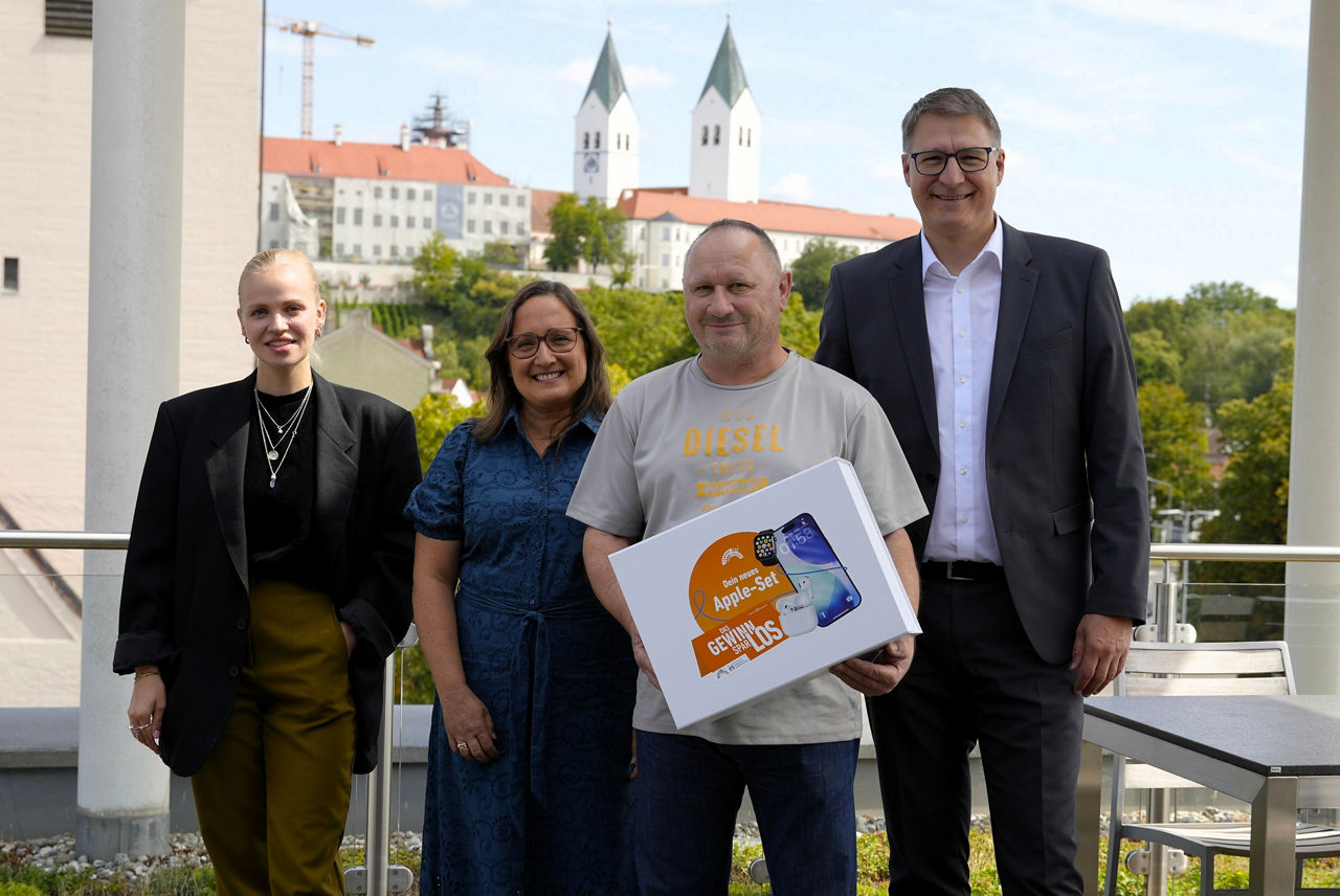 vier Personen stehen nebeneinander auf einer Terrasse. Ein Mann in hellem T-Shirt hält einen weißen Karton mit dem Apple-Set in Händen. Im Hintergrund die Dom-Silhouette Freisings