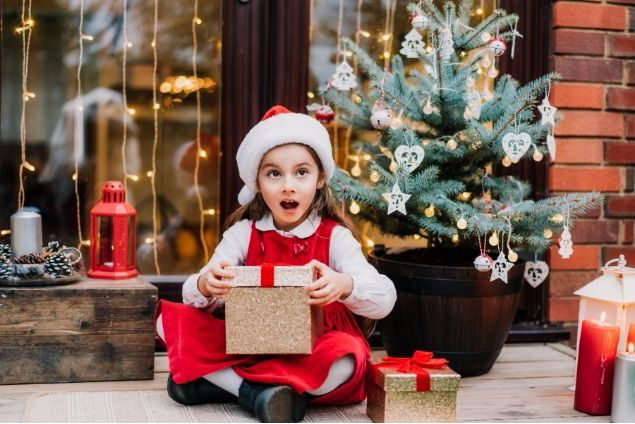 Kleines Mädchen mit Weihnchtsmannmütze packt ein Geschenk aus und schaut dabei überrascht