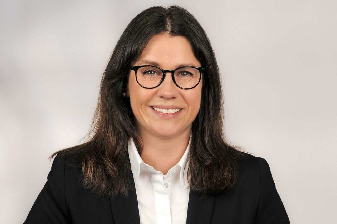 Simone Höflich hat dunkle mittellange Haare eine schwarze halbrunde Brille und trägt ein Businessoutfit