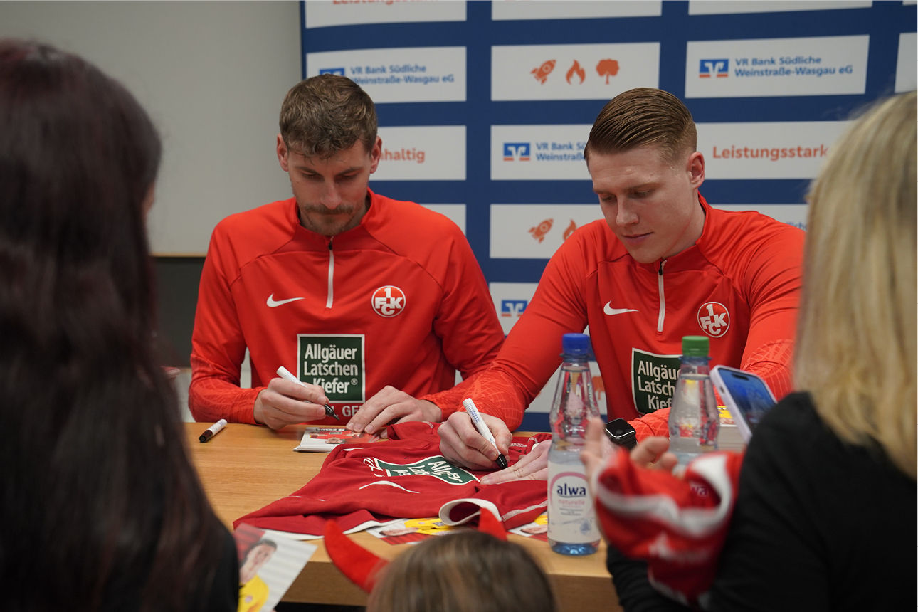 Zwei Fußballspieler signieren Trikots an einem Tisch