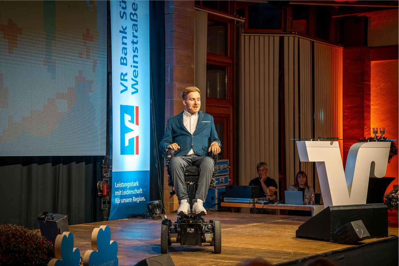 Mann im Anzug im E-Rollstuhl spricht auf Bühne bei Veranstaltung der VR Bank