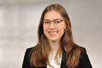 Frau mit langen, glatten Haaren, schwarzem Blazer und weißem Oberteil vor unscharfem Hintergrund, sie trägt eine Brille.
