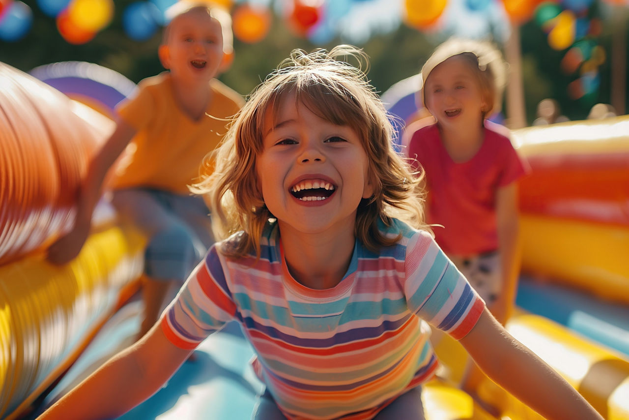 Auf einer bunten Hüpfburg sind drei lachende Kinder, eines im Vordergrund und zwei im Hintergrund.