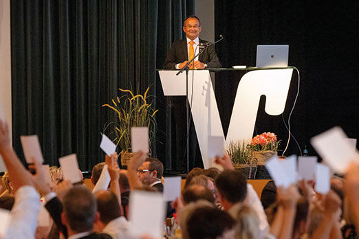 Mitglieder bei Versammlung heben Abstimmungskarten, Redner am Pult – demokratische Teilhabe und Engagement im Fokus.