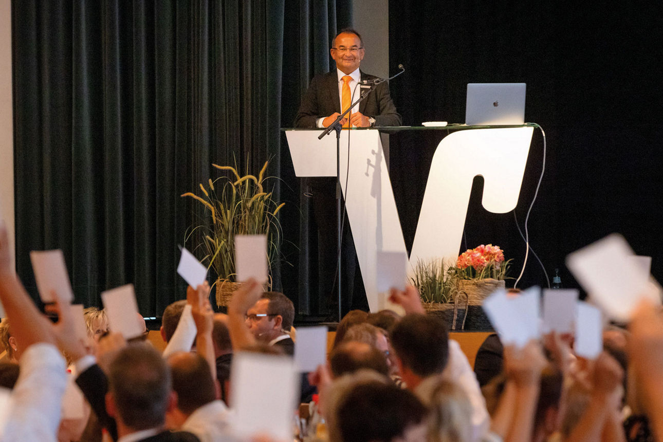 Mitglieder bei Versammlung heben Abstimmungskarten, Redner am Pult