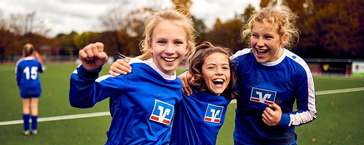 3 junge Fußballspielerinnen in Volksbank-Trikots stehen auf einem Fußballfeld