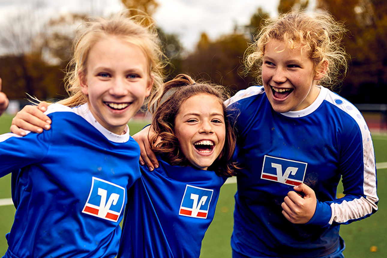 3 junge Fußballspielerinnen in Volksbank-Trikots stehen auf einem Fußballfeld