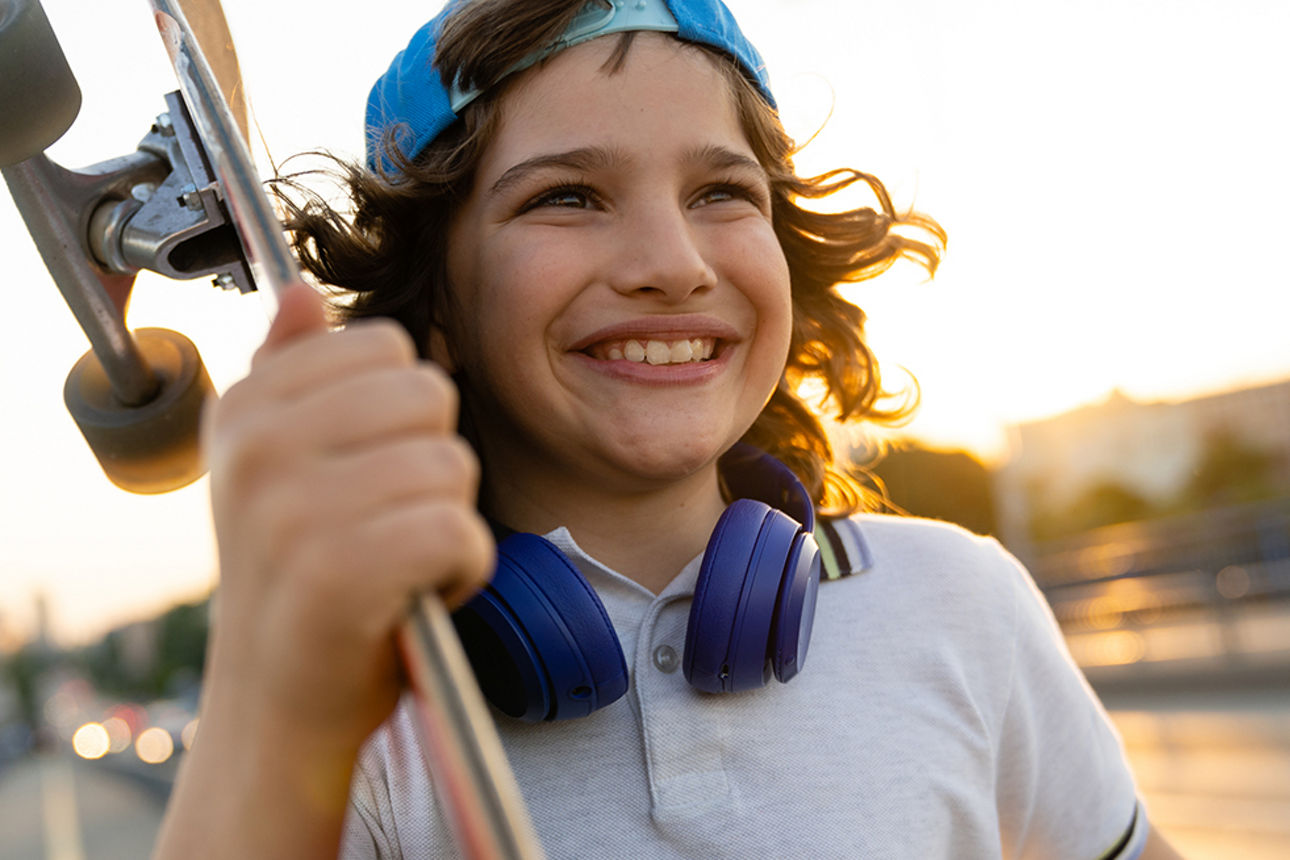 A boy is carrying his skateboard. Having fun on a summer evening. Boy is smiling while holding his skateboard. Skater boy wearing a white T-shirt and a blue baseball cap turned backwards.