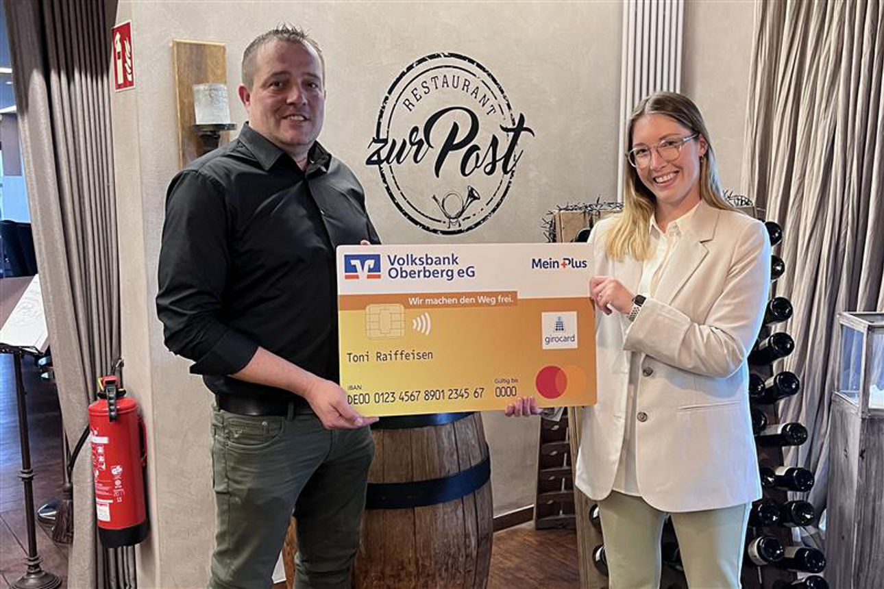 Ein Mann und eine Frau lächeln in die Kamera und halten gemeinsam eine übergroße, symbolische Bankkarte der Volksbank Oberberg.