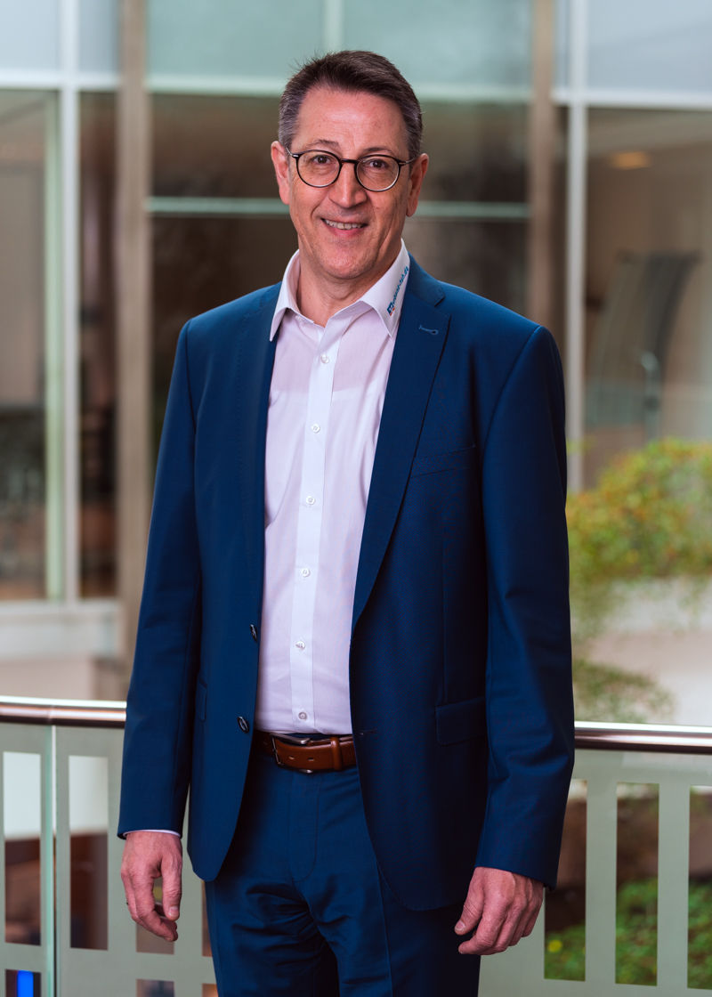 Ein Mann steht im dunklen Anzug vor einer Glaswand und lächelt in die Kamera.