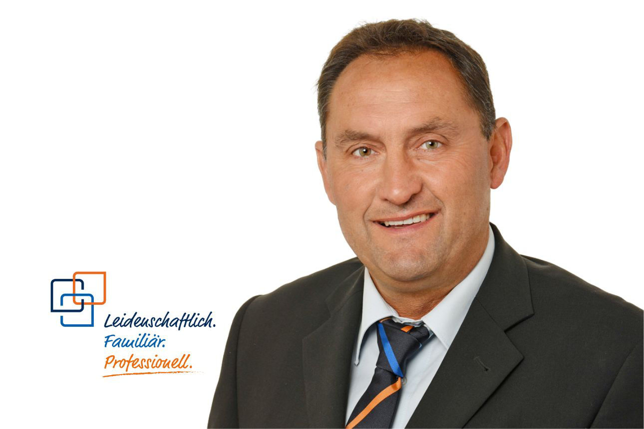 Porträt von Hans Harrer, einem lächelnden Mann mit kurzen dunklen Haaren im dunklen Anzug mit orange-blau gestreifter Krawatte. Daneben das Firmenlogo und der Slogan "Leidenschaftlich. Familiär. Professionell."