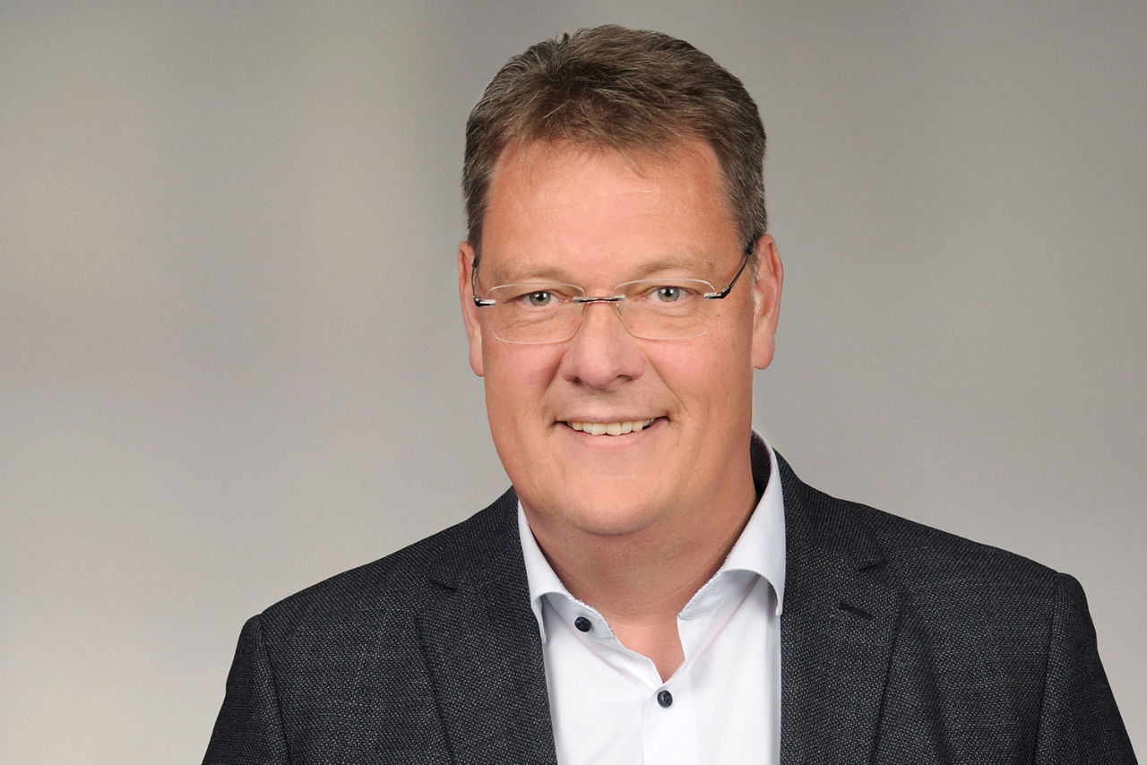 Mann mit kurzen braunen Haaren trägt weißen Hemd und dunklen Anzug vor neutralem Hintergrund