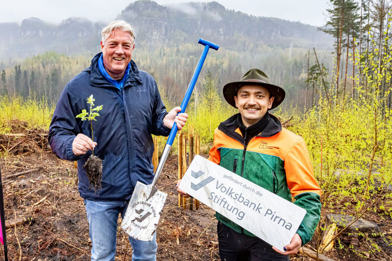 Zwei Männer in Regenkleidung grinsen in die Kamera, präsentieren stolz Setzling, Schild und Spaten