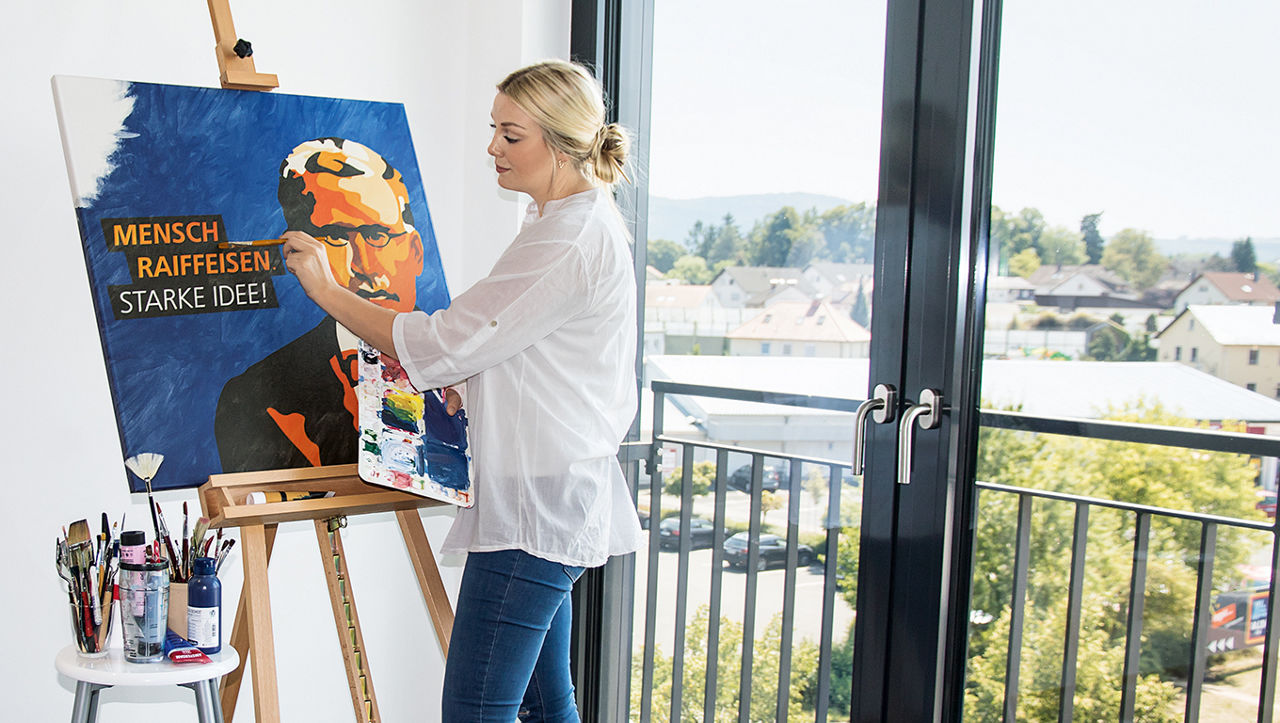 Eine Frau steht vor einer Staffelei und mal ein Bild von Friedrich Wilhelm Raiffeisen in orange und blau.