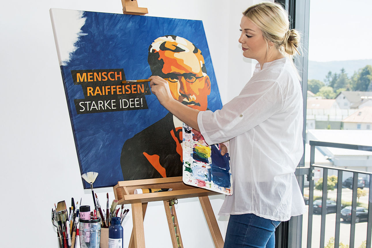 Eine Frau malt an einer Staffelei ein Porträt mit dem Text ‚Mensch Raiffeisen – Starke Idee‘. Sie steht in einem hellen Raum.