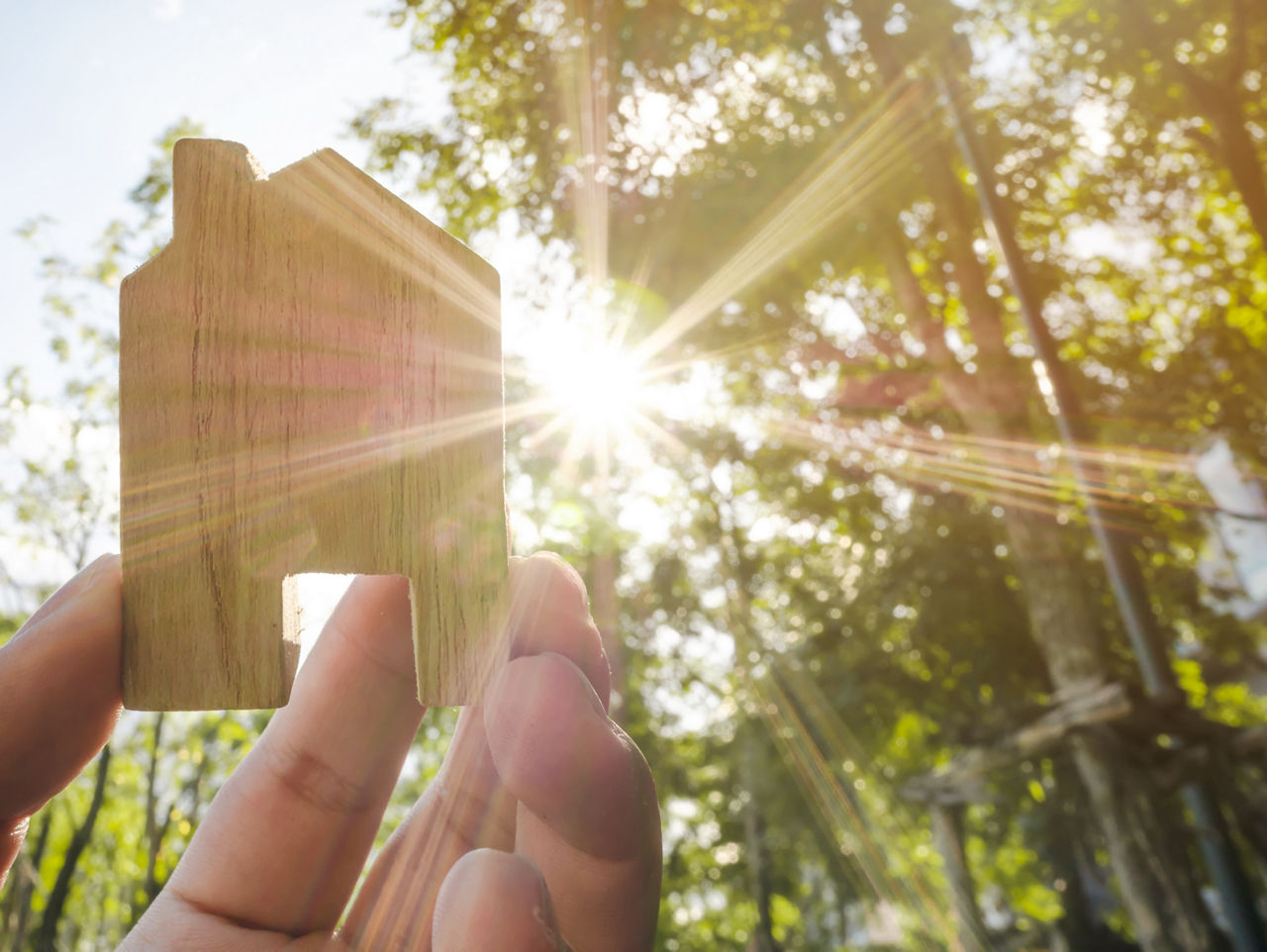 Hand hält ein kleines Holzhaus vor Sonnenlicht und Bäumen – Symbol für Nachhaltigkeit.