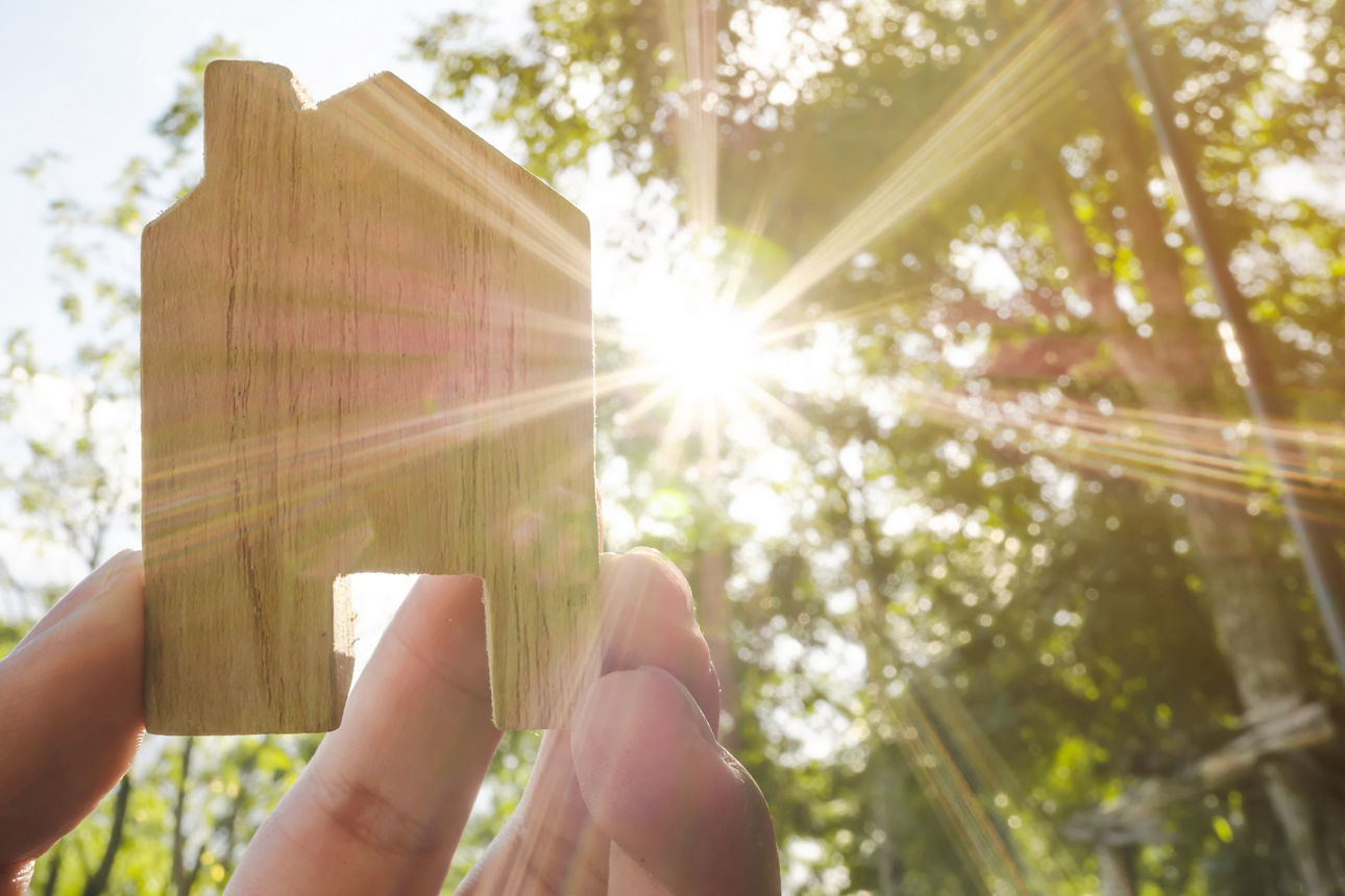 Hand hält ein kleines Holzhaus vor Sonnenlicht und Bäumen – Symbol für Nachhaltigkeit.