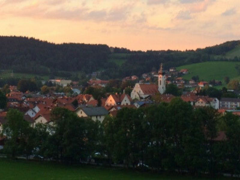 Eine panoramische Ansicht des Ortes Hausham bei Sonnenuntergang, mit Häusern, einer Kirche und bewaldeten Hügeln im Hintergrund.