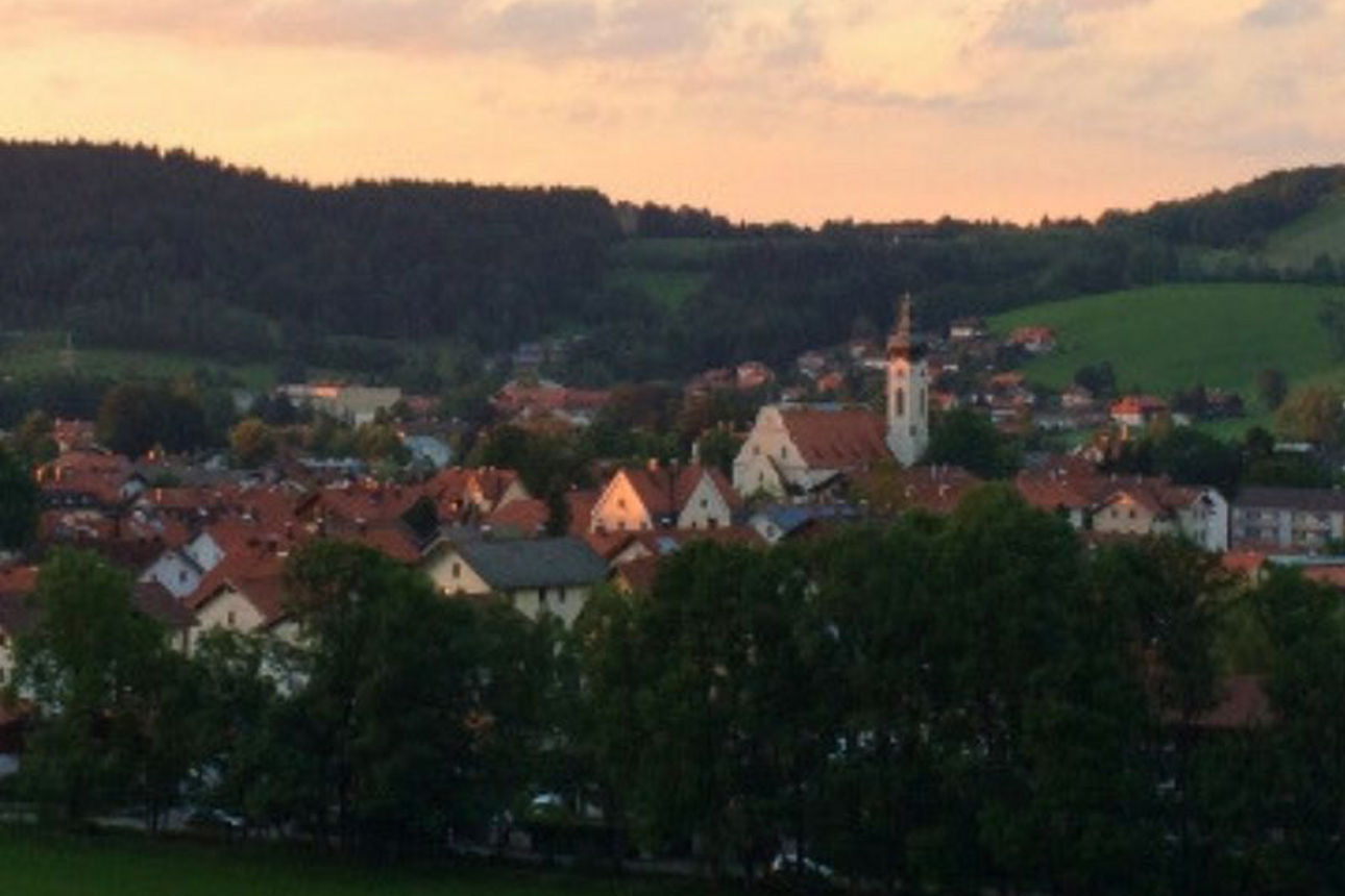 Eine panoramische Ansicht des Ortes Hausham bei Sonnenuntergang, mit Häusern, einer Kirche und bewaldeten Hügeln im Hintergrund.