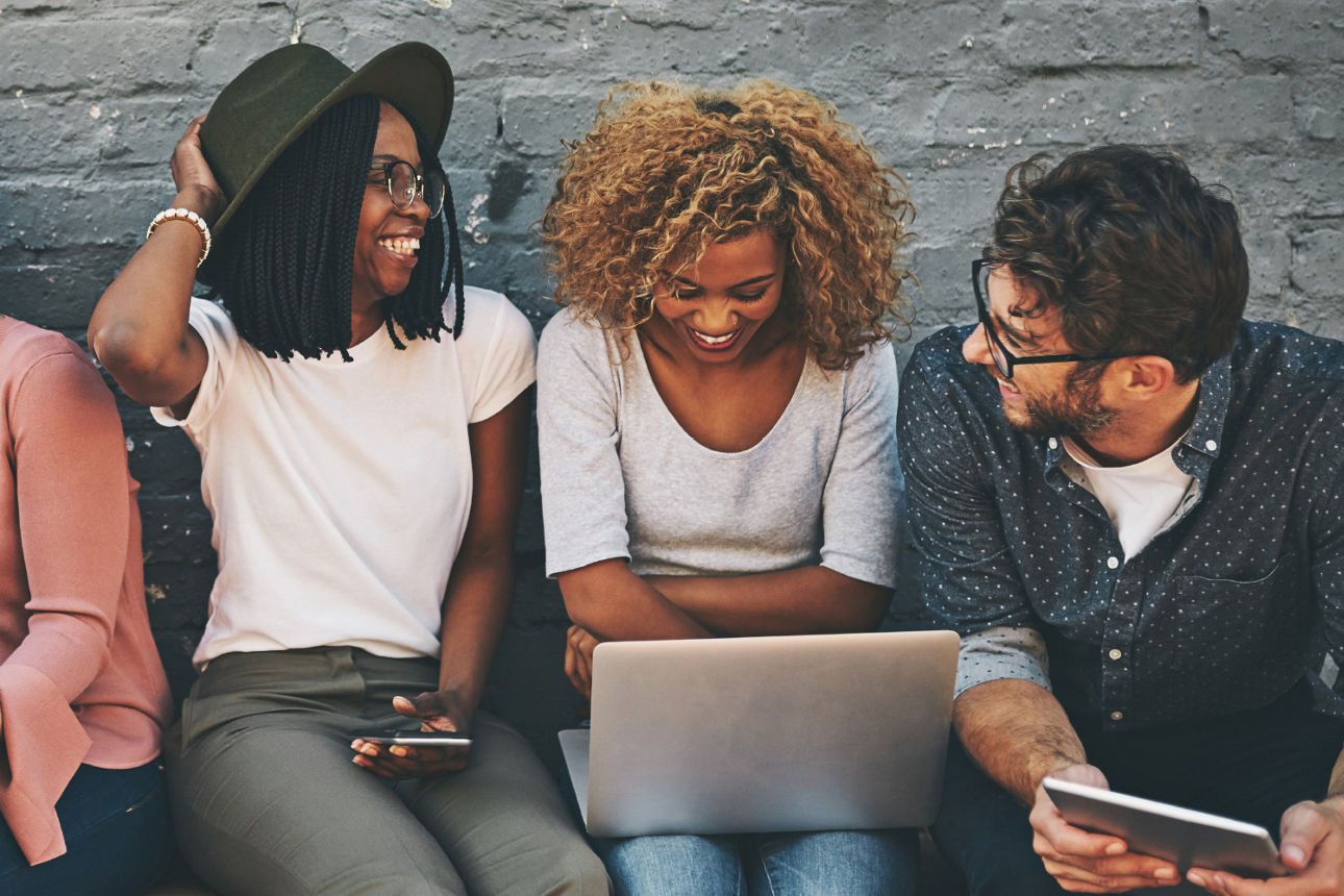 Mehrere Leute sitzen auf einer Bank und lesen auf verschiedenen Geräten (Laptop, Handy, Tablet)