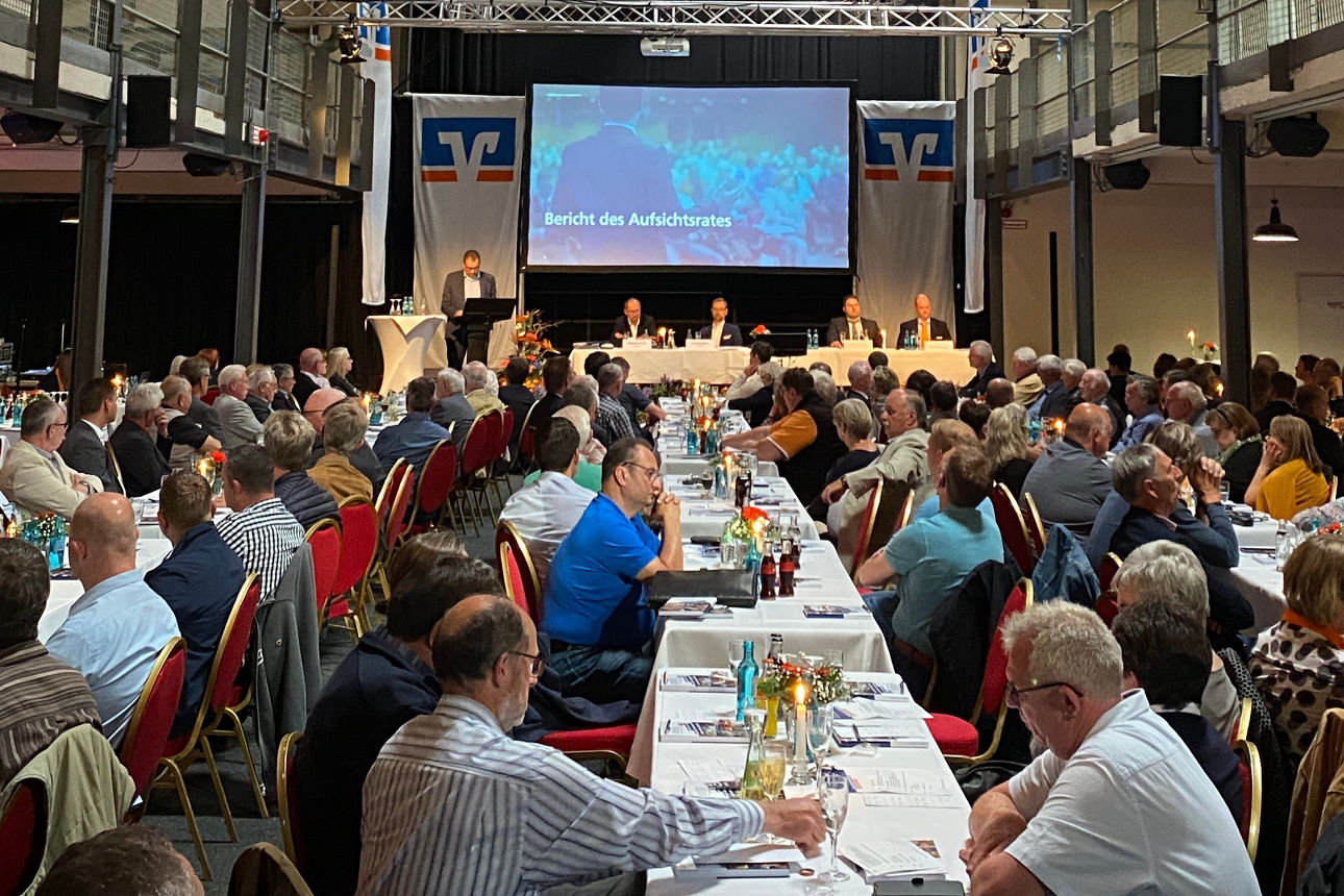 Mehrere Personen in formeller Kleidung sitzen und stehen in einem Veranstaltungsraum mit Bühne und Leinwand.